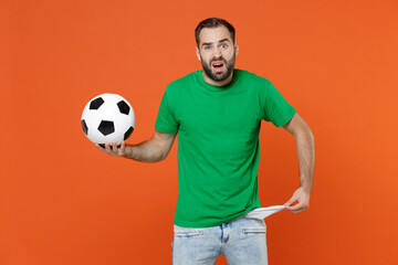 Wall Mural - Confused man football fan in green t-shirt cheer up support favorite team with soccer ball has no money showing empty pocket has no money isolated on orange background. People sport leisure concept.