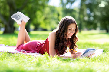 leisure, inspiration and people concept - happy smiling woman writing to diary or notebook lying on picnic blanket at summer park