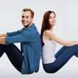 Happy amazed couple. Full body length profile portrait of sitting back to back models in love studio concept, isolated over bright grey background. Man and woman together. Square composition image.