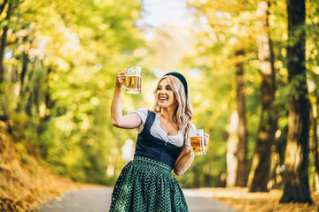 Wall Mural - Pretty happy blonde in dirndl, traditional festival dress, holding two mugs of beer outdoors in the forest