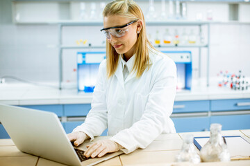 Female researcher in white lab coat and protective goggles using laptop while working in the laboratory