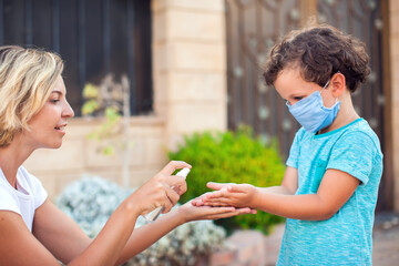 Wall Mural - Mother and kid with medical mask disinfecting hands with sanitizer outdoors. Life during coronavirus pandemic.