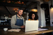© Prins Productions - Male and mixed race female coffee shop owners smiling at laptop in coffee shop.