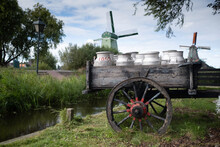 Milk Cans On An Old Wheel Cart In A Garden With Windmills In The Background In The Netherlands