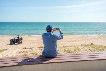 Wall Mural - Back view of man taking sea beach pictures by smart phone sunshine outdoors