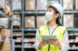 © ake1150 - Young asian woman auditor or trainee staff wears mask working during the COVID pandemic in store warehouse shipping industrial. looking up and checks the number of items store by digital tablet.