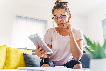 Wall Mural - Female student getting ready for exam reading on digital tablet sitting on sofa couch at home while studying in bright day wearing eyeglasses - education and leisure concept