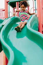 A Little Girl Sliding Down A Green Slide At The Playground.