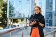 © Serhii - Successful female banker using tablet and wireless earphones outdoors near his office background lights, portrait young woman professional manager working on touch pad in evening city