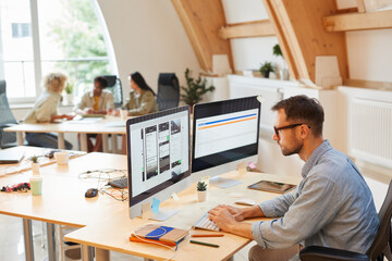Serious programmer sitting at his workplace using computers in his work he developing new software at office with his colleagues in the background