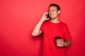 Portrait of a handsome young man in red t-shirt talk mobile phone and coffee cup isolated over red background