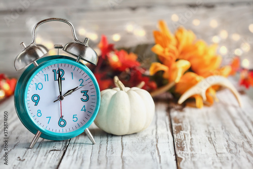 Set your clocks and fall back. Clock and decorations of mini pumpkins, colorful autumn leaves, antlers and bokeh lights over a white wooden table. Daylight saving time concept.