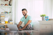 © My Ocean studio - Handsome young bearded man in casual clothes is working at home while sitting on the bed. Confident guy with a laptop and smartphone drinks coffee in bedroom.