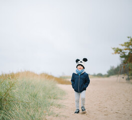 Smiling five years old boy wearing  jacket, scarf and hat walking  with his hands on pockets in the beach park