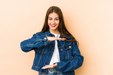 Young caucasian woman isolated en beige background holding something with both hands, product presentation.