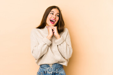 Young caucasian woman isolated en beige background keeps hands under chin, is looking happily aside.