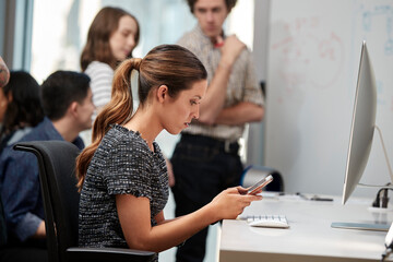 Young woman using cell phone at computer, co-workers having discussion around dry erase board in background 