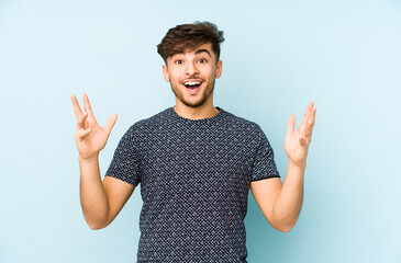 Wall Mural - Young arabian man isolated on a blue background receiving a pleasant surprise, excited and raising hands.