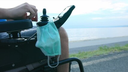 Wall Mural - Man putting face mask on wheelchair near sea at sunset. Protection from coronavirus.