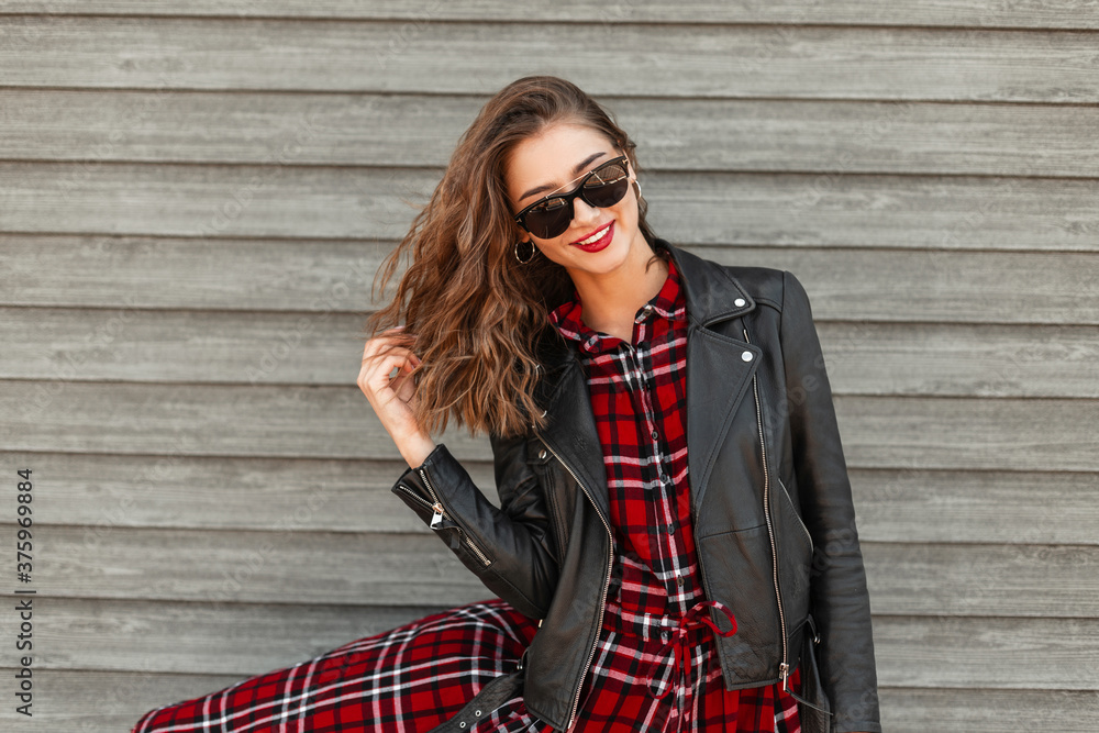Cute stylish joyful young woman with red lipstick with positive smile in sunglasses in fashion leather jacket near vintage wooden wall on street. Happy smiling woman model straightens hair in city. - obrazy, fototapety, plakaty 