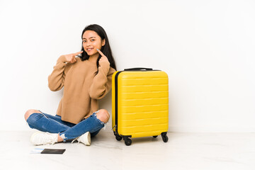 Young chinese traveler woman sittting on the floor with a suitcase isolated smiles, pointing fingers at mouth.