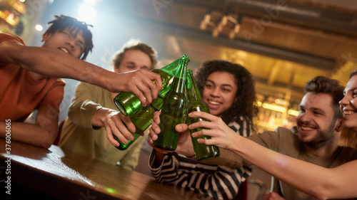 Fotomural A group of happy friends in the bar watching sports match on TV together, drinking beer, clinking bottles and cheering for team