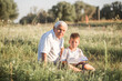 © Maria - Mid shot of grandfather and his grandson while reading a book together in meadow Small boy making first steps in read.