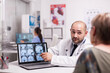 © DC Studio - Doctor discussing with senior woman with alzheimer in hospital office pointing at laptop screen with ct scan. Medic wearing white coat. Nurse in blue uniform working on computer.