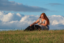 Plump Young Red-haired Blue-eyed Girl With A Labrador Retriever Photographed On A Field Against A Background Of Clouds.