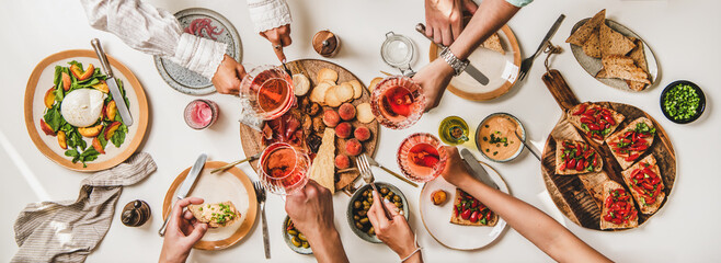 Wall Mural - Friends wine and snacks party. Flay-lay of people hands clinking glasses with rose wine over table with cheese, fruit, smoked meat, tomato brushettas, buratta salad, top view. Wine tasting concept