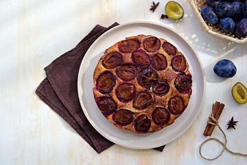 Wall Mural - Round honey cake or muffin with fresh plums on a light grater on a light concrete background. Plum baked goods.