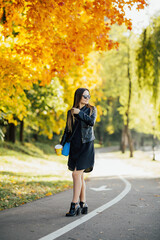 Stylish young woman with cup of coffee walking in autumn park. Woman in a black dress and a leather jacket  long hair wearing glasses with an empty road stretching into the distance. 