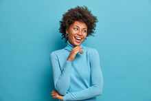 Portrait Of Dark Skinned Cheerful Woman With Curly Hair, Touches Chin Gently, Laughs Happily, Enjoys Day Off, Feels Happy And Enthusiastic, Hears Something Positive, Wears Casual Blue Turtleneck