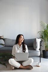 Vertical shot of creative young asian woman freelancer, working with laptop and looking outside window with dreamy smile. Student writing essay and thinking, sit on floor at home with computer