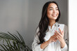 canvas print picture - Portrait of attractive modern asian woman looking outside window and smiling dreamy, holding smartphone. Girl waiting for phone call, working remote from home, planning schedule
