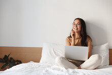 Work From Home, Freelance And Lifestyle Concept. Portrait Of Creative Young Asian Female Sitting On Bed With Laptop, Smiling Thoughtful And Looking Away While Think-up. Girl Shopping Online