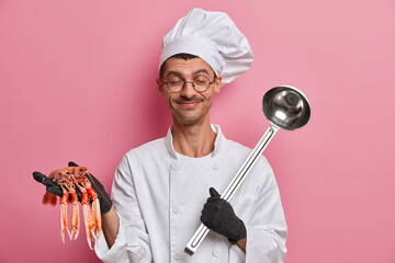 Horizontal shot of happy chef going to prepare seafood salad, holds crayfish, kitchenware, creats diet menu, tries new recipe, stands with closed eyes in special uniform. Culinary, kitchen concept