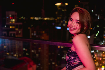 An attractive young asian woman poses by a rooftop bar with views of the city. At Makati, Metro Manila. Nightlife and dating in Manila.