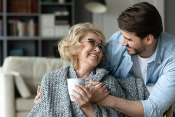 Wall Mural - Smiling mature woman holding cup, hugging with adult son close up, enjoying leisure time, happy elderly mother wearing warm scarf drinking hot tea or coffee, family spending weekend together at home