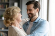 Close up smiling mature mother and adult son hugging, standing in living room at home, family enjoying tender moment, happy overjoyed beautiful middle aged woman and young man cuddling