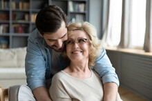 Happy Young Man Hugging Mature Mother Wearing Glasses From Back, Expressing Love And Gratitude, Adult Son And Middle Aged Woman Cuddling, Family Enjoying Tender Moment, Sitting In Living Room At Home