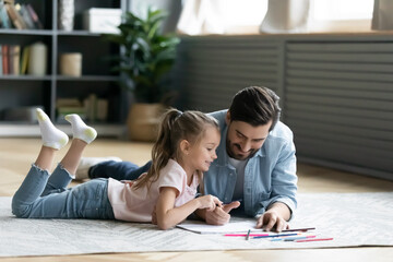 Loving father and adorable little daughter drawing with colored pencils, lying on warm floor with underfloor heating in modern living room, having fun together, family enjoying creative activity