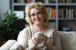 © fizkes - Head shot portrait smiling mature woman holding cup of tea sitting on cozy couch at home, positive overjoyed middle aged female wearing glasses looking at camera, enjoying free time at home