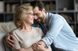 Smiling mature woman and adult son cuddling, enjoying tender moment, sitting on cozy couch in living room, happy beautiful elderly mother wearing glasses and young man hugging, two generations