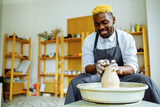Fototapeta Do przedpokoju - mixed race afro male potter with black apron sitting at workshop table potter