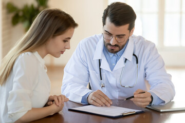 Doctor wearing white uniform and glasses consulting young woman about insurance contract terms or prescriptions, therapist gp and patient discussing medical checkup results at meeting