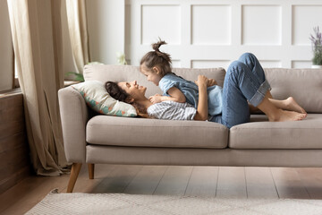 In cozy living room on comfy couch lying mother and her little cheery daughter. Kid girl lay on mom enjoy playtime and positive communication. Funny time together at home, love and protection concept