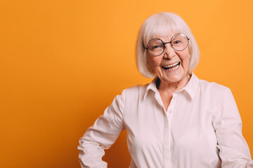 Image of happy old woman with light short hair wearing white blouse holding her arms on hips and smiling. Woman isolated over bright orange background.