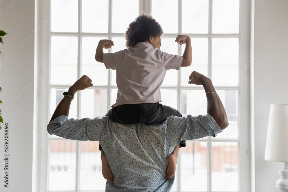Back side view young african american father holding on shoulders little preschool funny kid son, showing biceps. Fit strong different generations family enjoying domestic activity, healthcare concept - obrazy, fototapety, plakaty 