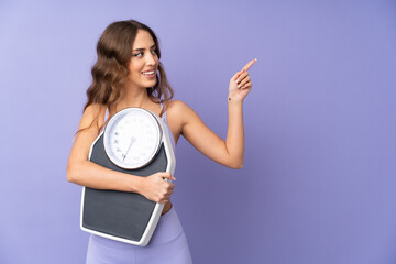 Young sport woman over isolated purple background with weighing machine and pointing side